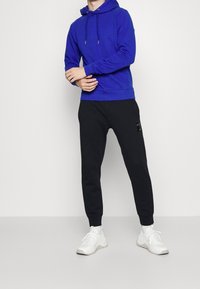 Uomo che indossa un felpa blu con cappuccio, pantaloni jogger neri con una toppa laterale logo e scarpe da ginnastica bianche, in piedi su uno sfondo bianco uniforme.