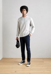 Gray sweatshirt with ribbed cuffs, paired with navy pants and black sneakers. Model holds a small black bag. Wood floor background.
