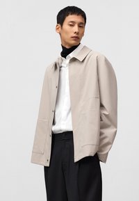 Lichtbeige jas met een klassieke kraag, knoopsluiting en grote zijzakken, gedragen over een wit shirt en zwarte broek. Zachte textuur.