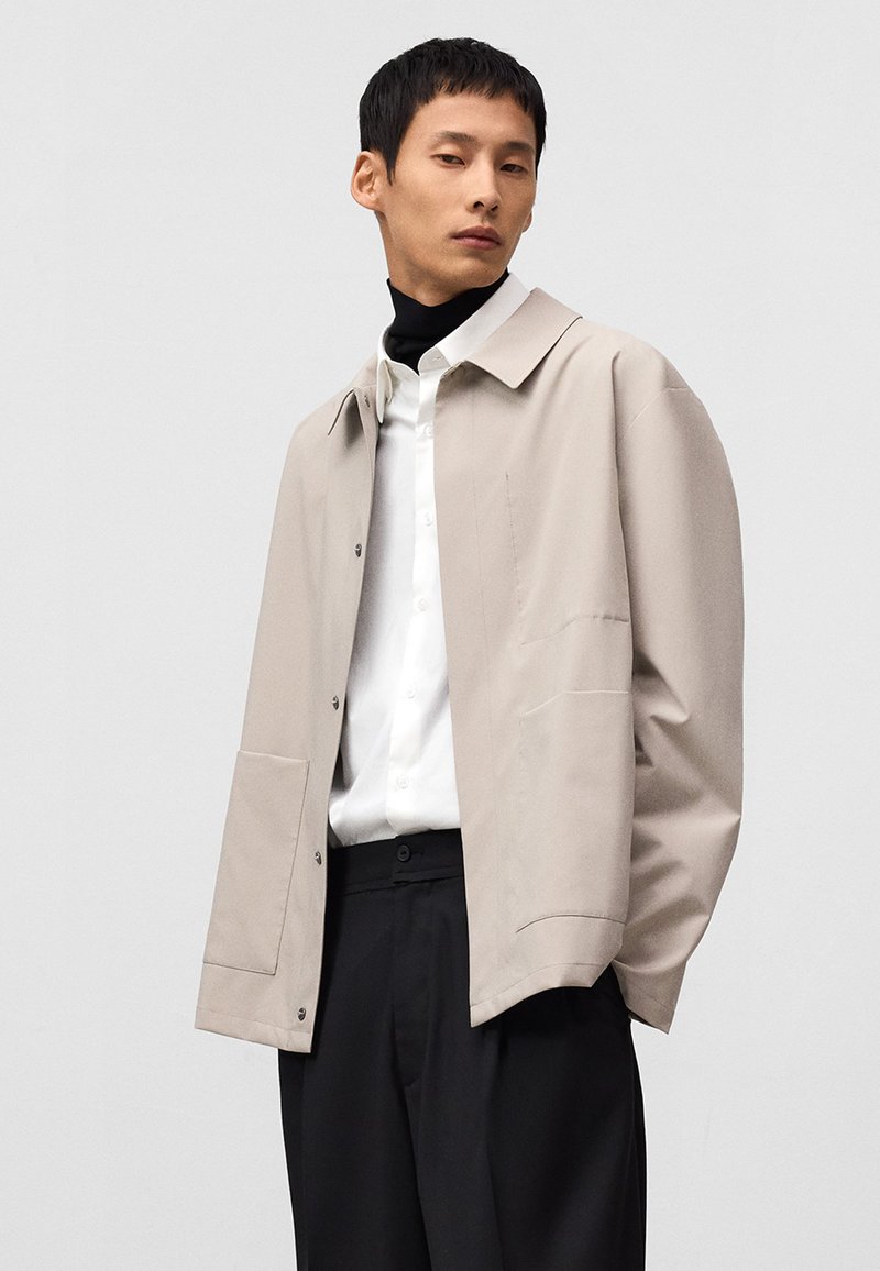 Lichtbeige jas met een klassieke kraag, knoopsluiting en grote zijzakken, gedragen over een wit shirt en zwarte broek. Zachte textuur.