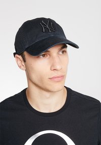 Gorra de béisbol negra con logotipo bordado, visera curvada y correa de tela en la parte posterior. Llevada con una camiseta negra simple.