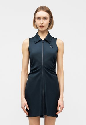 Kvinde med kort lyst hår iført en ærmeløs sort kjole med lynlås foran og krave mod en ensartet lys baggrund.