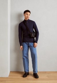 Suéter azul marino de cremallera, jeans desgastados y botas negras de tobillo. La modelo lleva un pequeño bolso cruzado con un diseño rectangular. Suelo de madera, pared gris claro.