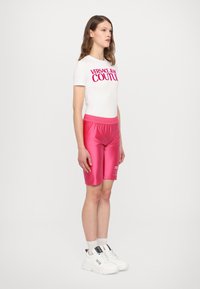 Bílá košile s krátkými rukávy a růžovým logem "VERSACE JEAN COUTURE", ve spojení s lesklými růžovými šortkami a bílými teniskami, které mají texturovaný pas.