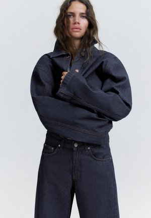 Donna con lunghi capelli mossi che indossa una giacca di jeans scura oversize abbinata a jeans a vita alta, in piedi davanti a uno sfondo chiaro e neutro.