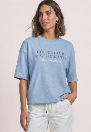 Vrouw draagt lichtblauw shirt met korte mouwen met tekst "Coffee Club New York City" en witte broek, één hand in de zak, kijkt iets naar rechts.