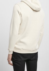 Sweat à capuche de couleur crème avec une texture douce, doté d'une capuche à cordon de serrage et de poignets côtelés. Porté avec un jean noir.