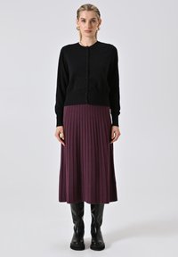 Frau trägt goldene Creolen, einen schwarzen knöpfbaren Cardigan, einen lila plissierten Midirock und schwarze kniehohe Stiefel und steht vor einem einfarbigen Hintergrund.