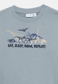 Blå barneskjorte med ulike dinosaurillustrasjoner over teksten "SPIS, SOV, BRUL, GJENTAR!" i fet mørke bokstaver.