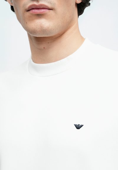 Άσπρο μπλουζάκι με στρογγυλή λαιμόκοψη που διαθέτει ένα μικρό κεντητό μαύρο λογότυπο στο κέντρο. Ομαλή ύφανση με εφαρμοστό σχέδιο.