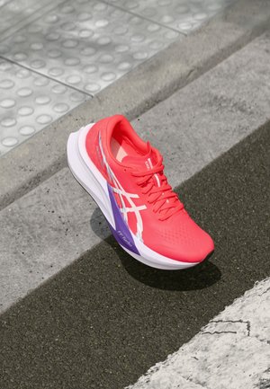 Lyserød løbesko med hvide striber og lilla detaljer på mellemsålen, hvilende på kantstenen og taktil belægning.