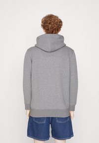 Grauer Hoodie mit einer großen, doppelt genähten Kapuze, aus weichem Stoff gefertigt. Gerippte Bündchen und Saum. Getragen mit Jeansshorts. Rückenansicht.
