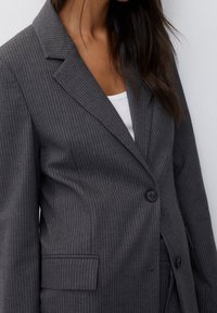 Blazer gris à fines rayures avec un revers cranté, une fermeture à un bouton et des poches latérales. Le tissu semble texturé avec une silhouette structurée.