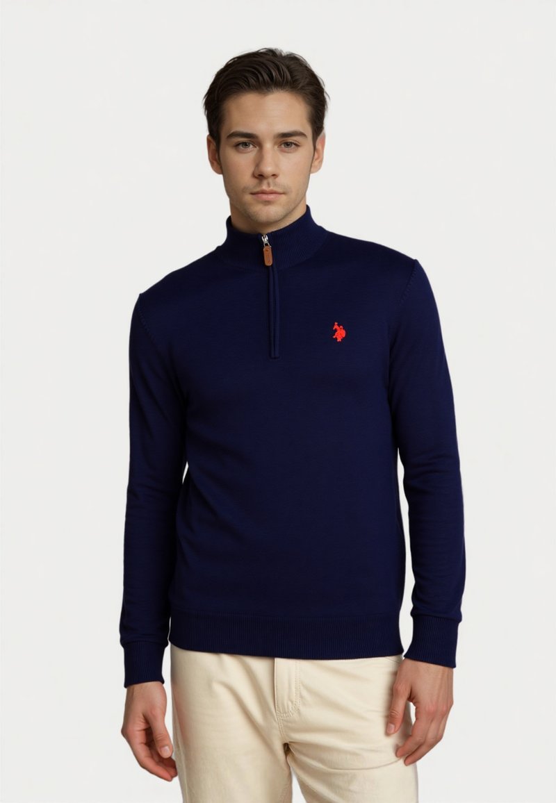 Junger Mann, der einen navyblauen Quarter-Zip-Pullover mit einem roten gestickten Logo und beigen Worten vor einem weißen Hintergrund trägt.