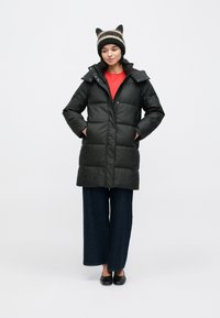 Čierny prešívaný kabát so zipsom a zapínaním na gombíky, kapucňou a dvoma bočnými vreckami, v kombinácii s červenou košeľou, nohavicami s širokými nohavicami a pletenou čiapkou.