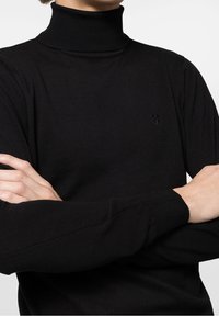 Pull à col roulé noir avec texture côtelée. Manches longues, coupe ajustée. Petit logo discret sur la poitrine. Tissu lisse.