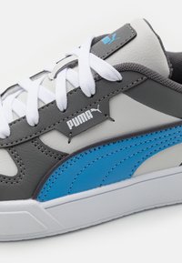 Grå och vit sneaker med blå accenter, med en texturerad läderöverdel, vita snören och en logotyp på sidan. Gummisula och sömdetaljer.