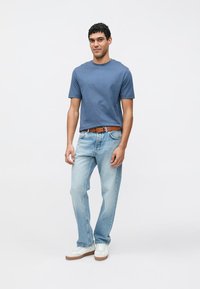 Modra bombažna majica, svetlo modre kavbojke, rjav pas in bele superge z rjavimi poudarki. Model stoji na nevtralnem ozadju.