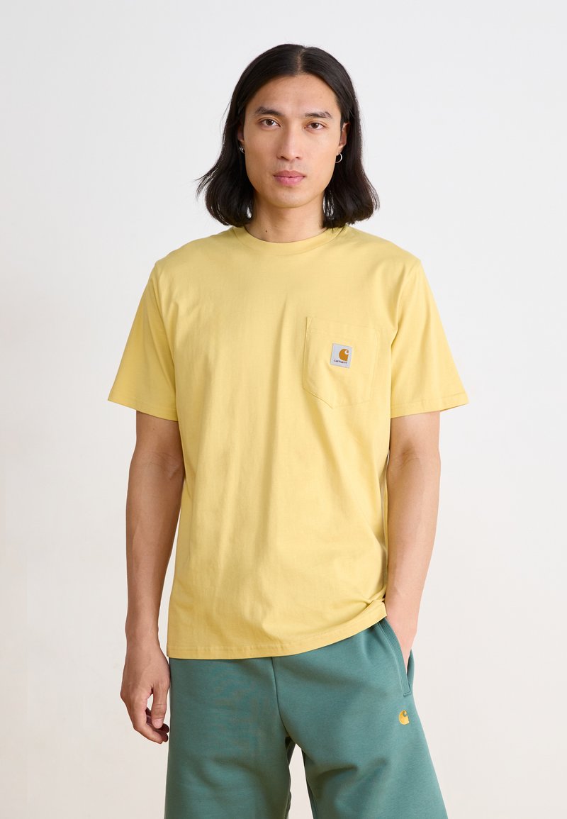 T-shirt en coton jaune à manches courtes avec une poche poitrine et un petit logo. Associé à un pantalon de survêtement vert, mettant en avant une coupe décontractée et un look casual.