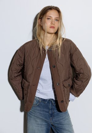 Jeune femme aux longs cheveux blonds portant une veste matelassée marron, une chemise blanche et un jean bleu sur un fond clair uni.