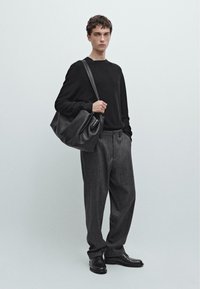 Maglione nero, pantaloni grigi su misura e scarpe nere. Questa persona tiene una grande borsa di pelle morbida con una lunga tracolla.