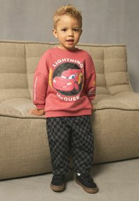 Un jeune enfant se tenant à côté d'un canapé beige, portant un sweat-shirt rouge de Lightning McQueen et un pantalon à carreaux noir et gris avec des chaussures noires.