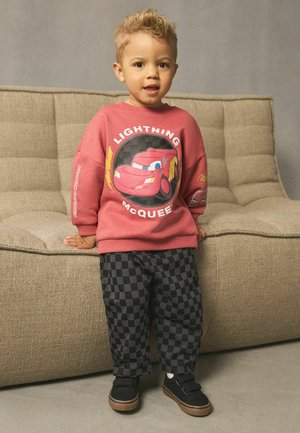 Ein junges Kind steht neben einem beigen Sofa und trägt einen roten Lightning McQueen-Pullover sowie schwarz-grau karierte Hosen und schwarze Schuhe.