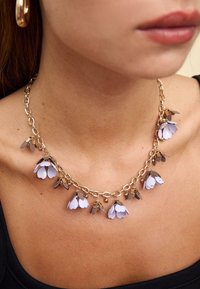 Zlati verižni ovratnik z lila in rjavimi cvetnimi poudarki; vključuje kovinske povezave in majhne sferične detajle.