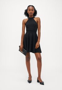 Robe noire à encolure halter avec un corsage ajusté et une jupe évasée. Associée à des chaussures noires à talons épais et à une pochette noire structurée.