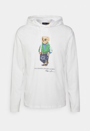 Biała bluza z kapturem z długim rękawem z grafiką niedźwiedzia w okularach przeciwsłonecznych, zielonej kurtce, pasiastym swetrze i granatowych spodniach, oznaczona jako Polo Bear by Ralph Lauren.