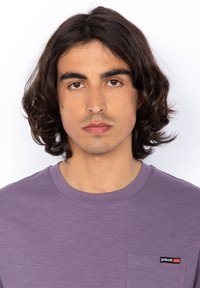 Una maglietta a maniche corte viola presenta una tasca e un piccolo logo Schott NYC rosso e nero. Il tessuto sembra liscio con una leggera texture.