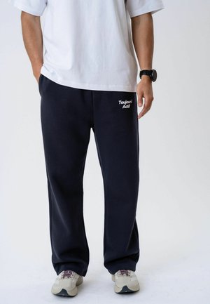 Personne portant un pantalon de jogging noir avec le texte « Toujours Actif », un t-shirt blanc, des baskets beiges et une montre noire au poignet, debout devant un fond uni.