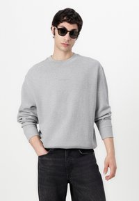 Gray waffle-knit sweatshirt with dropped shoulders, ribbed cuffs, and a subtle logo detail on the chest. Paired with dark denim jeans.