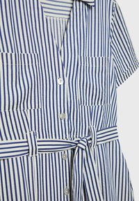 Robe à manches courtes à rayures verticales bleues et blanches avec poches avant, fermeture par boutons, col et ceinture assortie en tissu nouée à la taille.