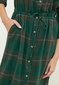 Abito a quadri verde realizzato in tessuto morbido, con maniche corte, vita con cordoncino e bottoni in legno lungo la parte frontale. Tasche laterali visibili.