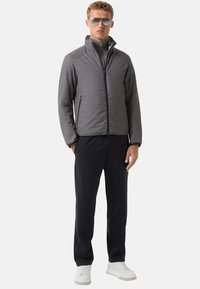 Graue, Reißverschlussjacke mit hohem Kragen, aus einem strukturierten Material, kombiniert mit dunklen Hosen und weißen Turnschuhen. Flache visuelle Details.