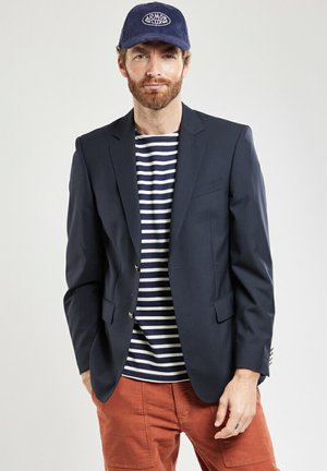 Homme avec barbe portant un blazer marine, une chemise rayée bleu marine et blanche, un pantalon couleur rouille et une casquette marine avec le logo Armor Lux.