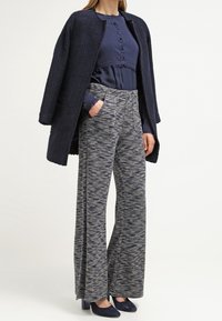 En marinblå texturerad överrock över en blå blus, kombinerad med mönstrade grå vida byxor och mörkblå pumps. Helhetsstil som blandar toner och mönster.