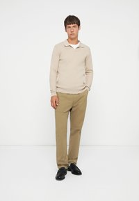 Beige geribbelde longsleeve polo, witte binnenlaag, gecombineerd met lichtbruine broek en zwarte schoenen. Eenvoudige, op maat gemaakte silhouet.