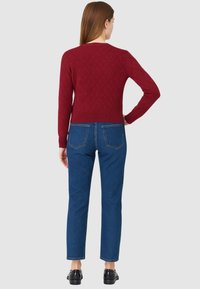 Donna in piedi di spalle, che indossa un maglione rosso con texture, jeans blu a gamba dritta e mocassini neri, su uno sfondo neutro.