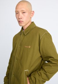 Veste vert olive avec un col, des boutons sur le devant et des détails de couture blancs. Présente le logo Timberland sur la poitrine à gauche.