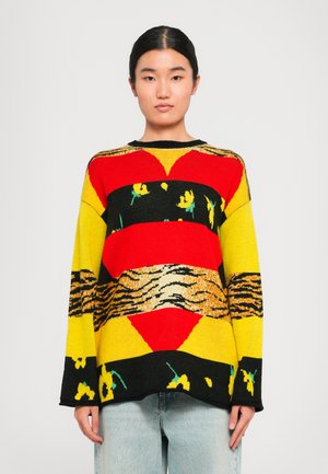 Woman wearing a colorful knit sweater with red, yellow, black, floral, and tiger stripe patterns, paired with light blue jeans.