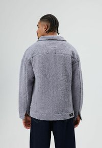 Grijze, zachte fleecejas met kraag, verlaagde schouders en een licht oversized pasvorm. Voorzien van een klein logopatch aan de zoom.