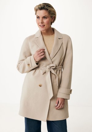 Beige Wollmischungsjacke mit Gürtel in der Taille, Reverskragen, doppelten Knöpfen und Ärmelakzenten. Getragen über einem leichten Strickoberteil und Jeans.