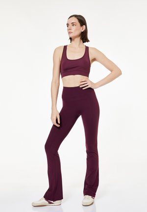 Vrouw staat met hand op heup, draagt een bijpassende donkerpaarse sportbeha en hoge, uitlopende legging met witte sneakers.