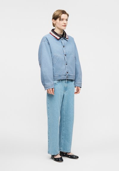 Άτομο που στέκεται, κοιτώντας μπροστά, φοράει ανοιχτό μπλε τζιν μπουφάν με γιακά με σχέδια, ανοιχτό μπλε τζιν παντελόνι και μαύρα πάνινα παπούτσια με διακοσμητικά.