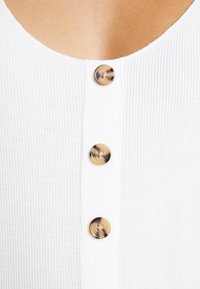 Bílý pletený top s třemi kulatými, želvovinovými knoflíky vpředu. Materiál má texturovaný povrch.