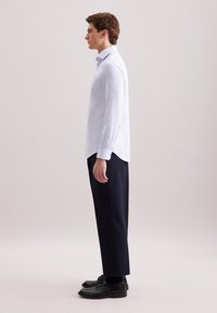 Camicia a quadri blu chiaro con maniche lunghe, dotata di colletto button-down e spacchi laterali; abbinata a pantaloni scuri e scarpe nere.