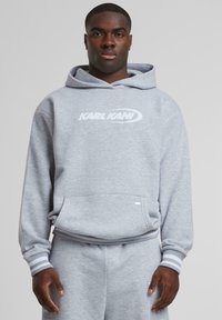 Gray hoodie made of soft fabric with a front pocket. Features a white logo "KARL KANI" and ribbed cuffs. Styled with matching gray sweatpants.