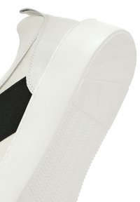 Baskets blanches avec une tige en cuir lisse, insert élastique noir et semelle en caoutchouc avec un motif de bande de roulement texturé.